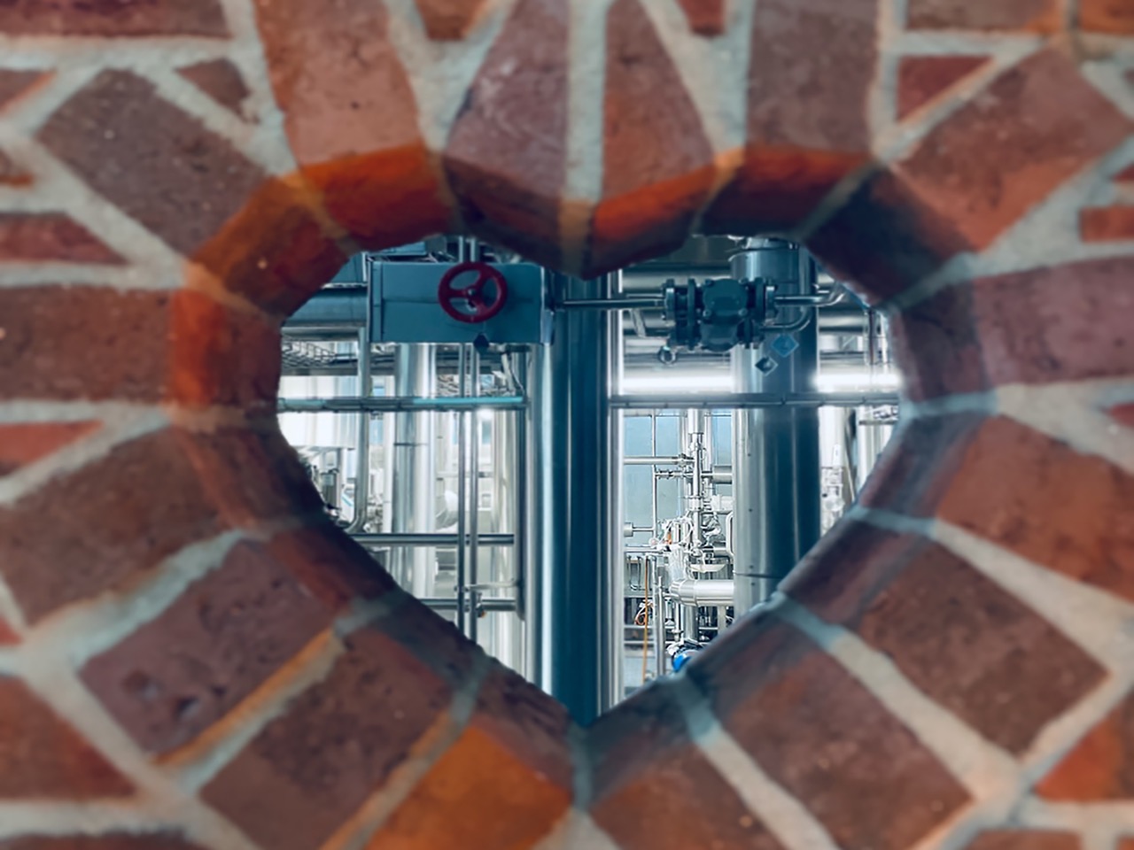 Herzförmiger Blick durch einen Ziegelsteinrahmen auf industrielle Anlagen im Hintergrund.