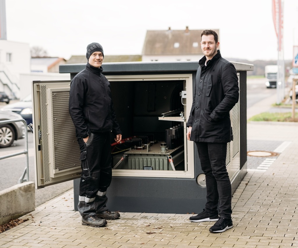 Zwei Männer stehen vor einem geöffneten technischen Schrank auf der Straße, umgeben von urbaner Umgebung.