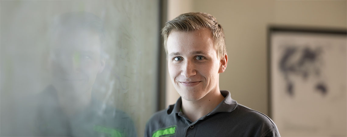 Ein junger Mann mit kurzem, blondem Haar lächelt vor einer transparenten Oberfläche, die einen schemenhaften Hintergrund zeigt.