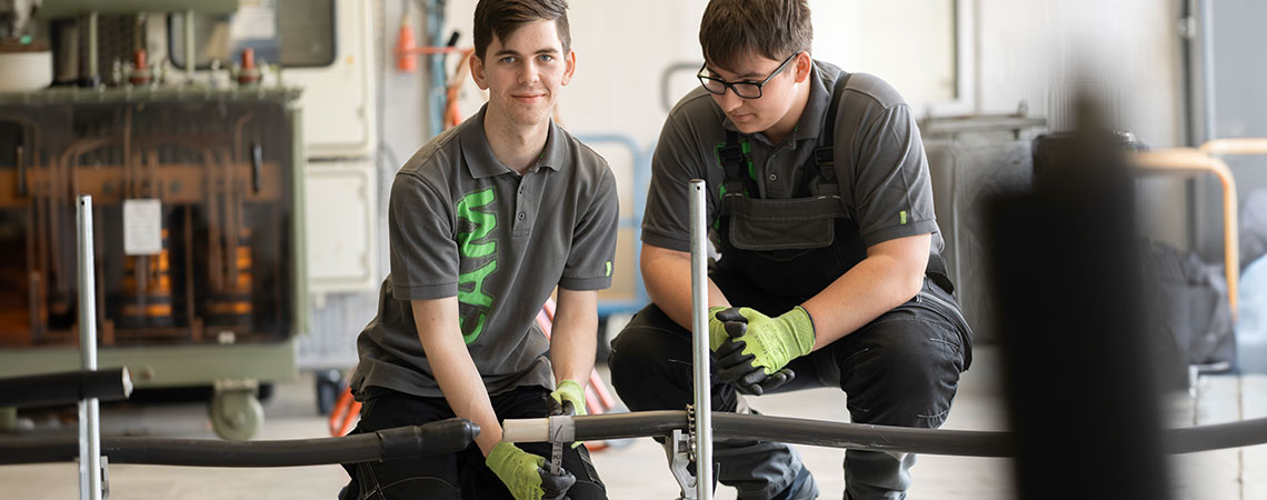 Zwei junge Männer arbeiten in einer Werkstatt an einem technischen Projekt mit einem Rohr und Werkzeug.