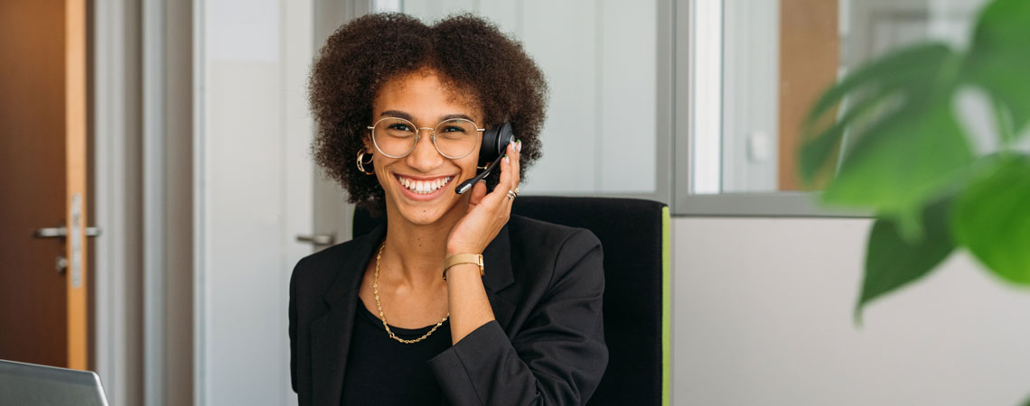 Lächelnde Person mit Headset in einem modernen Büro, sitzt vor einem Laptop und spricht am Telefon.