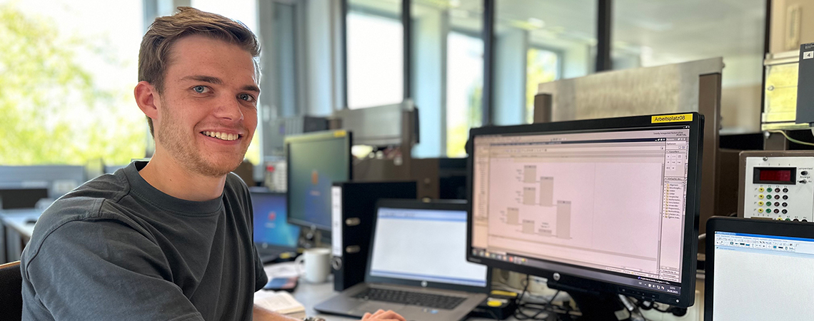 Junger Mann lächelt am Schreibtisch vor mehreren Monitoren und arbeitet an einem Computerprojekt.