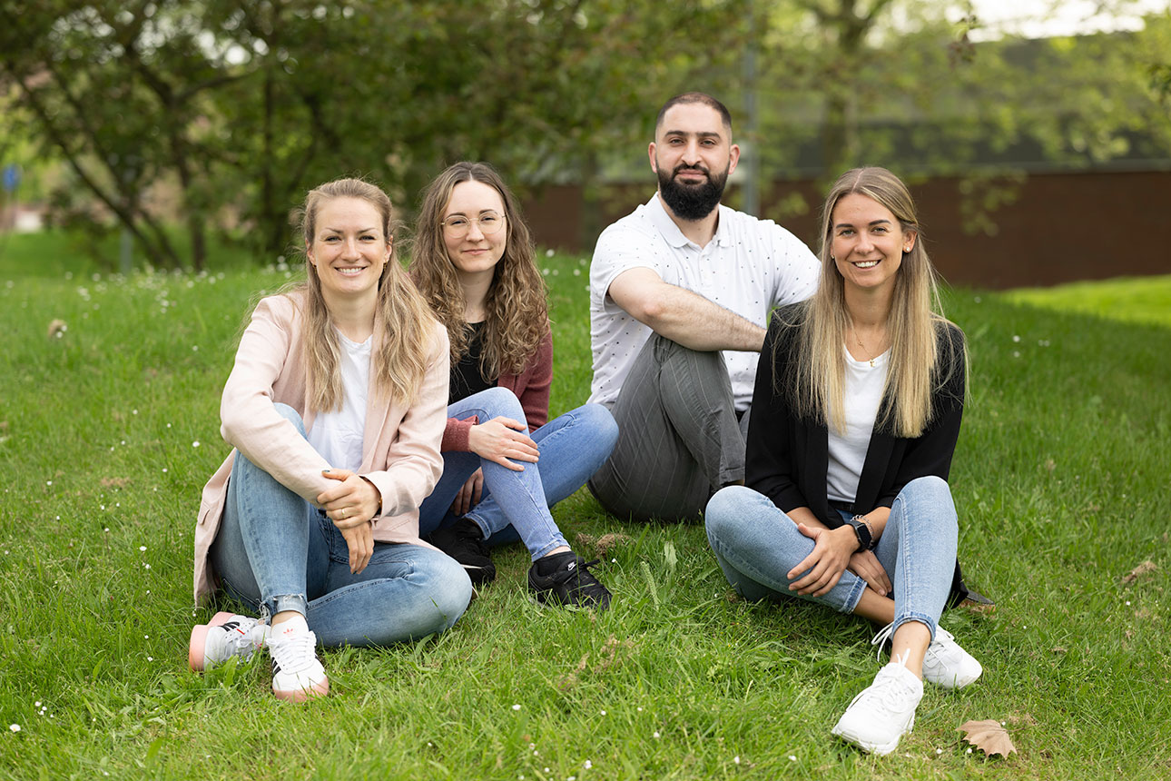 Vier Personen sitzen lächelnd auf einer grünen Wiese, umgeben von Bäumen und Blumen.