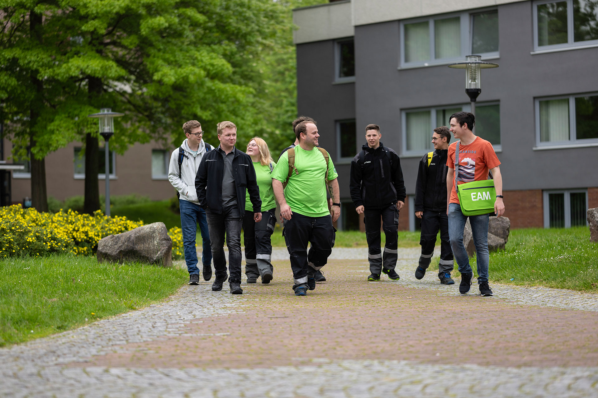 Sechs Personen gehen auf einem Weg, umgeben von Gras und gelben Blumen, vor einem modernen Gebäude.