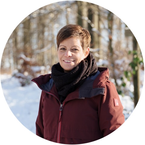 Frau mit kurzer brauner Haarfrisur in warmer Jacke und schwarzem Schal, steht lächelnd im Schnee.
