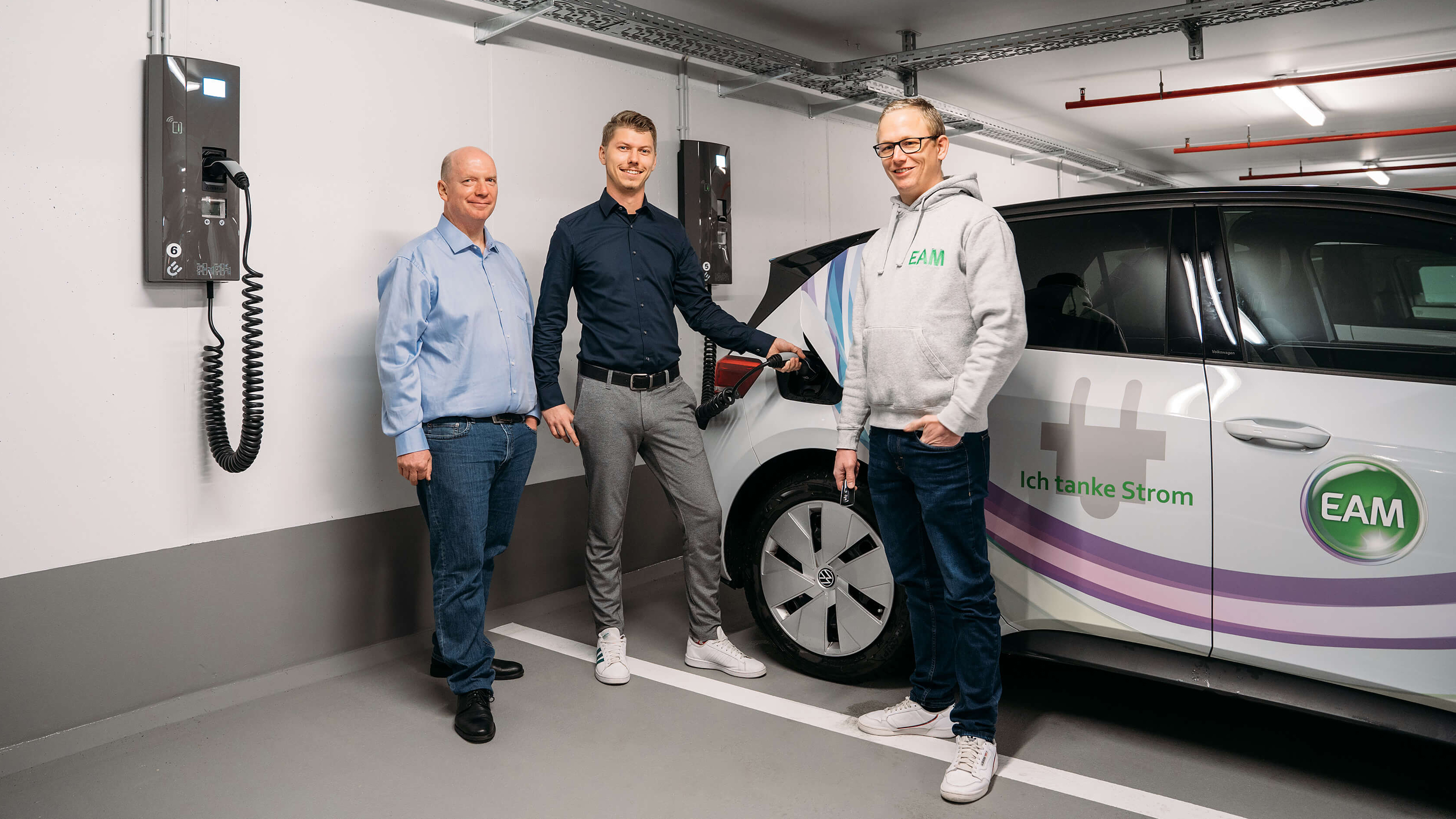 Drei Männer stehen an einer Ladestation für Elektroautos und halten ein Ladekabel in der Hand.