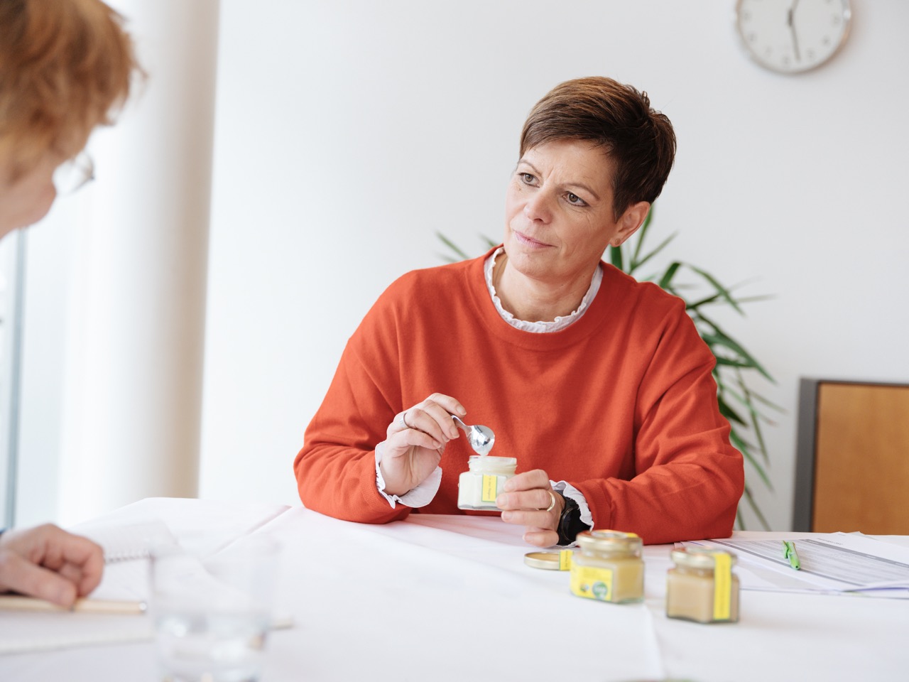 Eine Frau in orangefarbener Kleidung hält einen Löffel über einem Glas mit Speise und schaut interessiert.