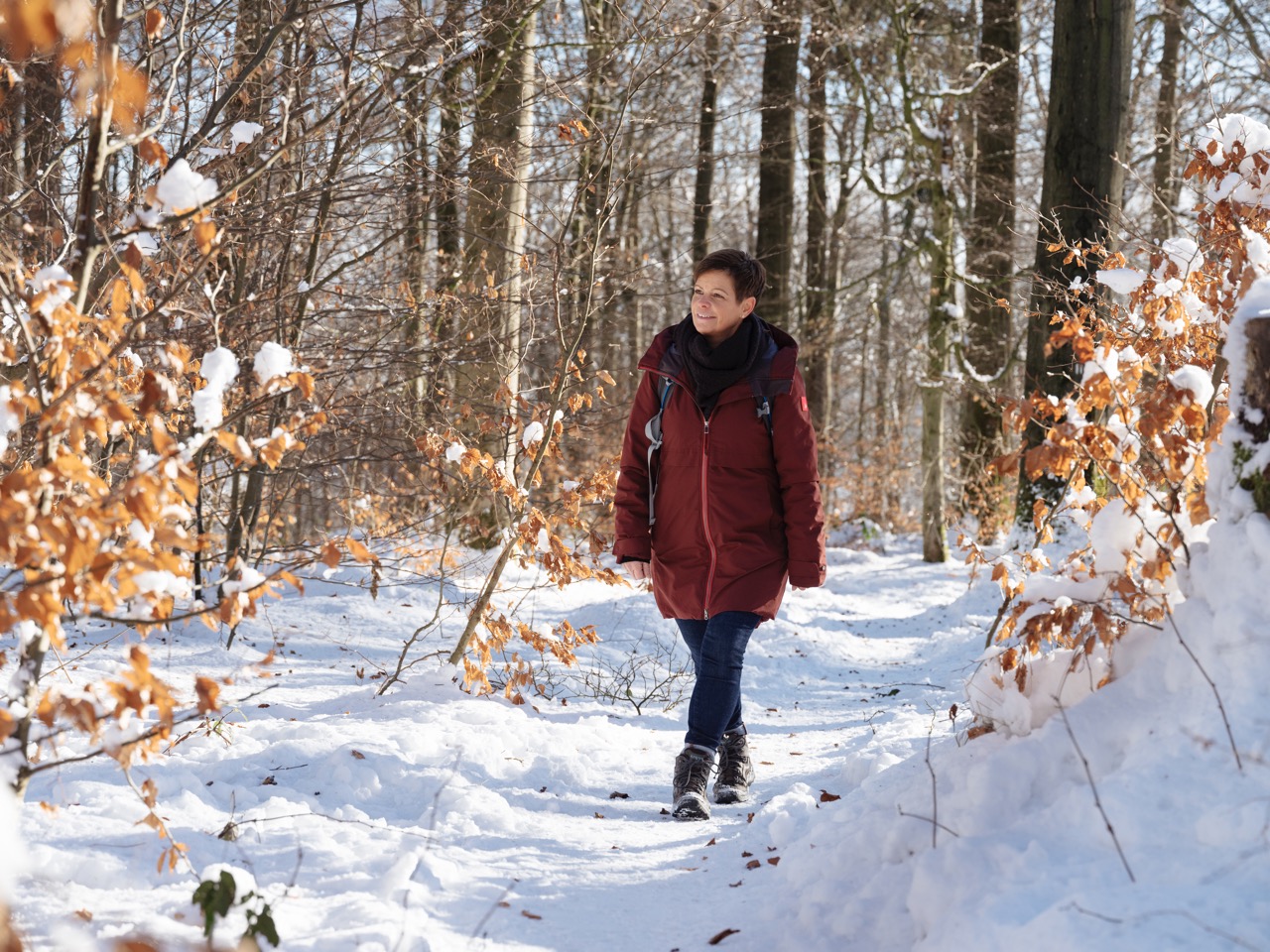 Eine Person in roter Jacke geht auf einem schneebedeckten Weg durch einen winterlichen Wald mit Laubbäumen und Schnee.