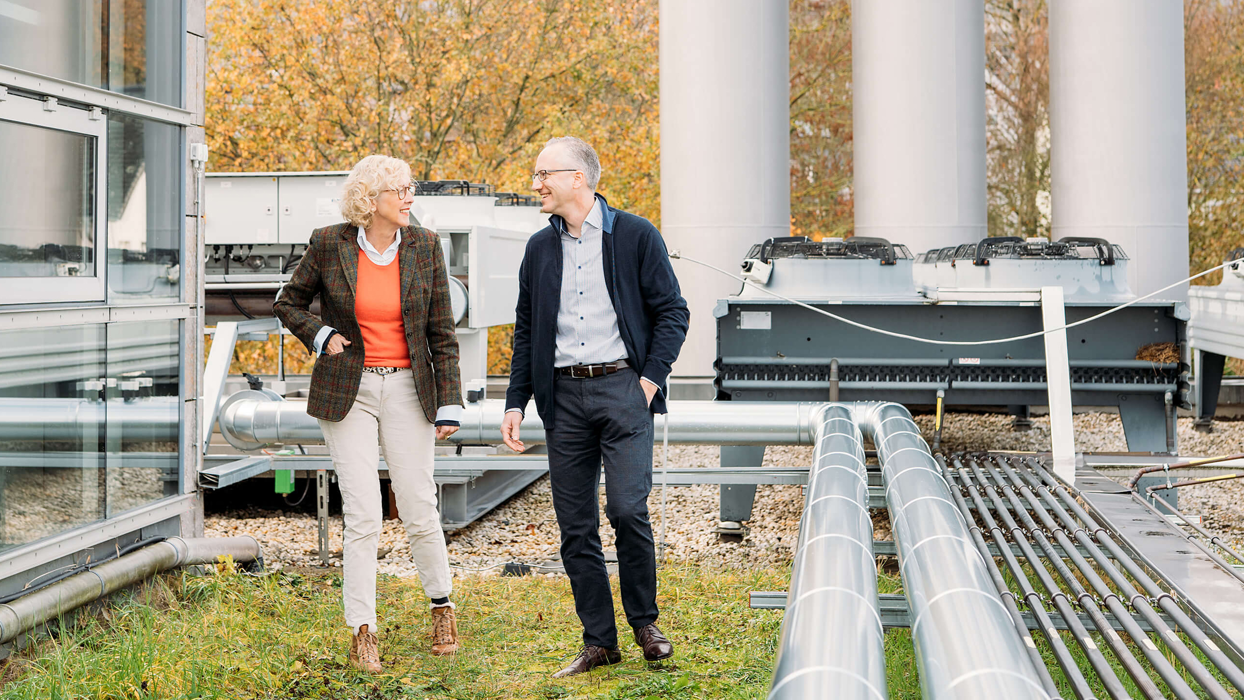 Zwei Personen spazieren auf einem Gelände mit technischen Anlagen, umgeben von grüner Wiese und Bäumen im Hintergrund.