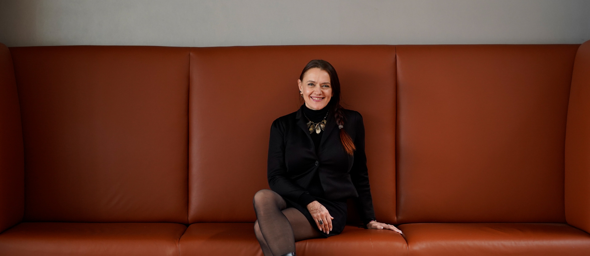 Frau in schwarzer Kleidung sitzt lächelnd auf einer braunen Couch, graue Wand im Hintergrund.