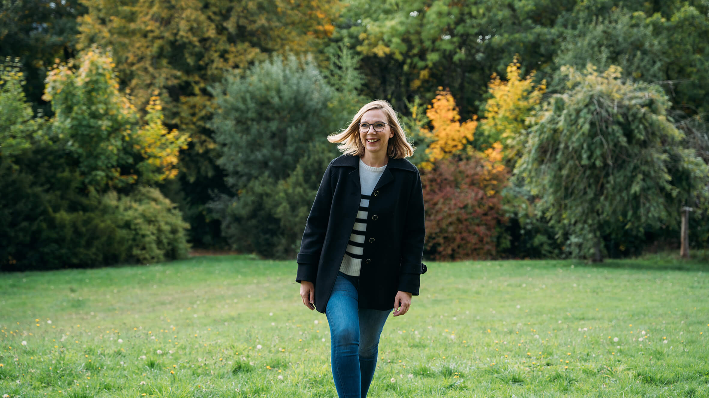 Eine lächelnde Frau mit Brille, die in einem grünen Park mit bunten Bäumen spaziert. Sie trägt eine schwarze Jacke und Jeans.