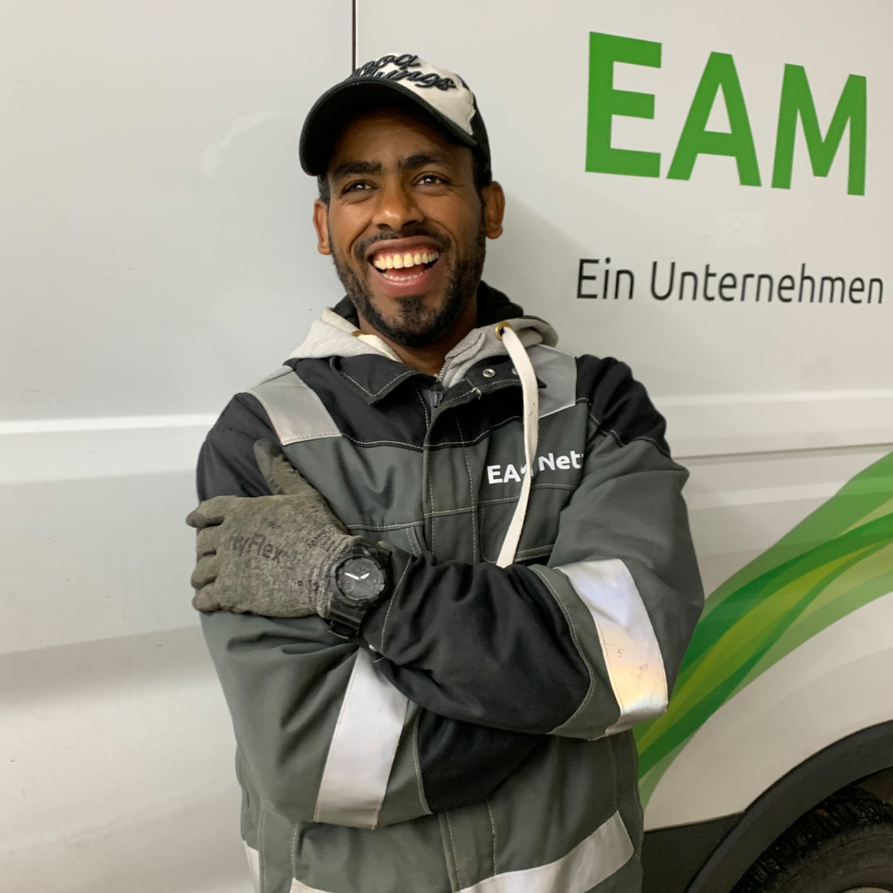 Lächelnder Mann in Arbeitskleidung mit Handschuhen steht vor einem Fahrzeug mit dem Schriftzug "EAM""."