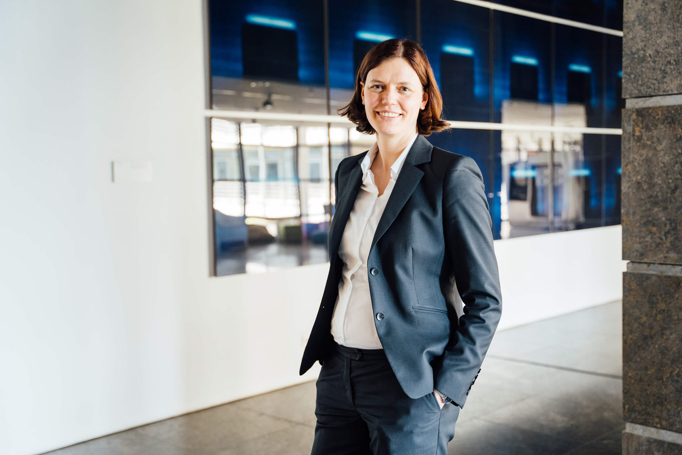 Eine lächelnde Frau in einem dunkelgrauen Anzug steht in einem modernen Büro mit großen Fenstern und digitalen Displays im Hintergrund.