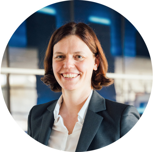 Porträt einer lächelnden Frau mit dunklem Haar, die einen grauen Blazer trägt, vor einem modernen Hintergrund.