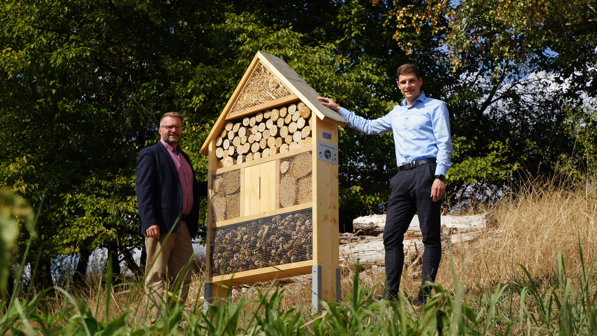 Zwei Männer stehen neben einem Insektenhotel in einer grünen Wiese, umgeben von Bäumen und hohem Gras.