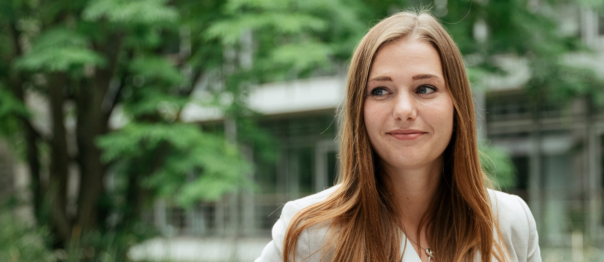Lächelnde Frau mit langen, braunen Haaren, im Freien vor grünen Bäumen, in einem freundlichen Moment.