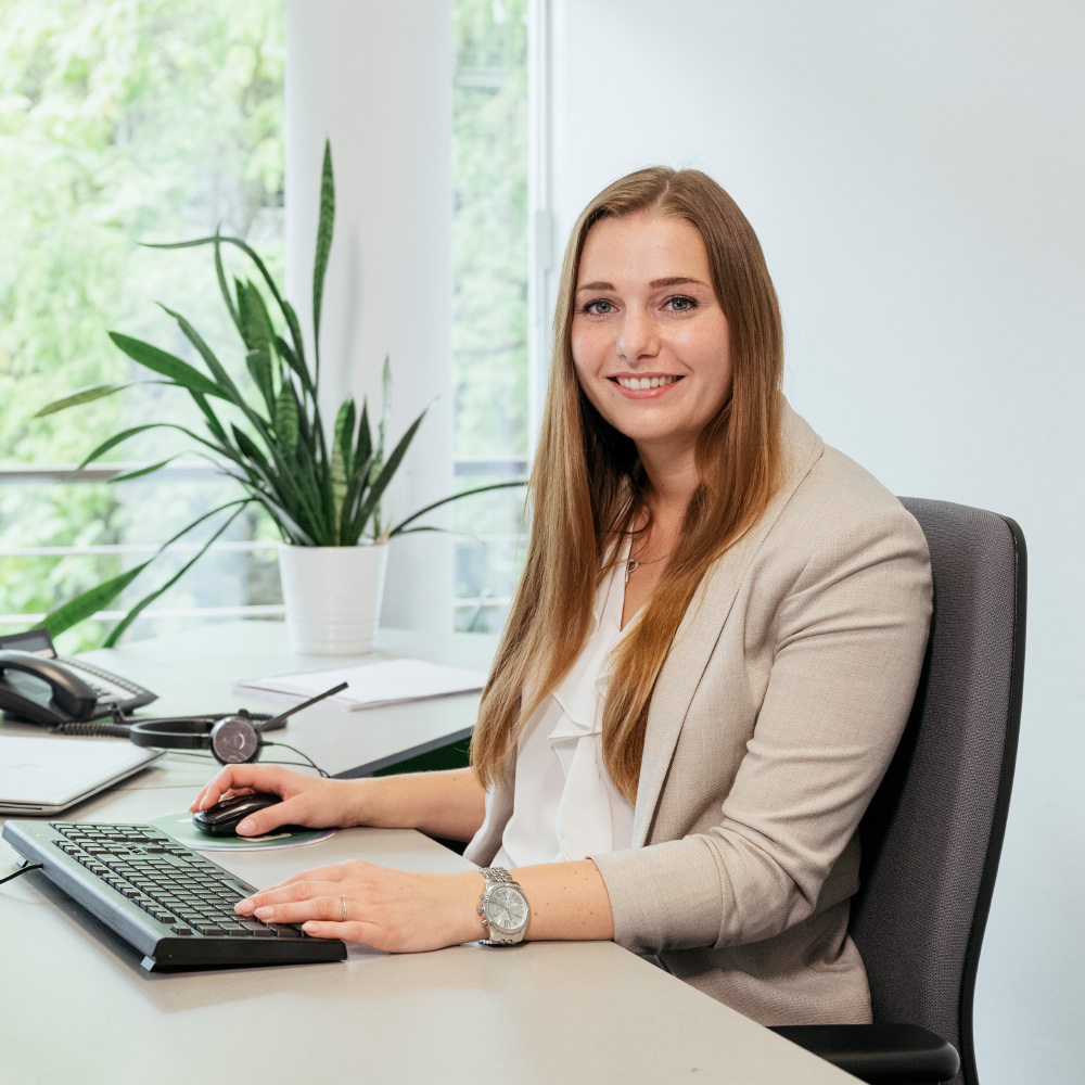 Frau mit langen Haaren in einem Büro, lächelt, sitzt an einem Schreibtisch mit Tastatur und Pflanzen im Hintergrund.
