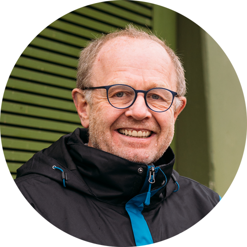 Mann mit Brille und lächelndem Gesicht, trägt eine schwarze Jacke mit blauen Akzenten, vor einer grünen Wand.