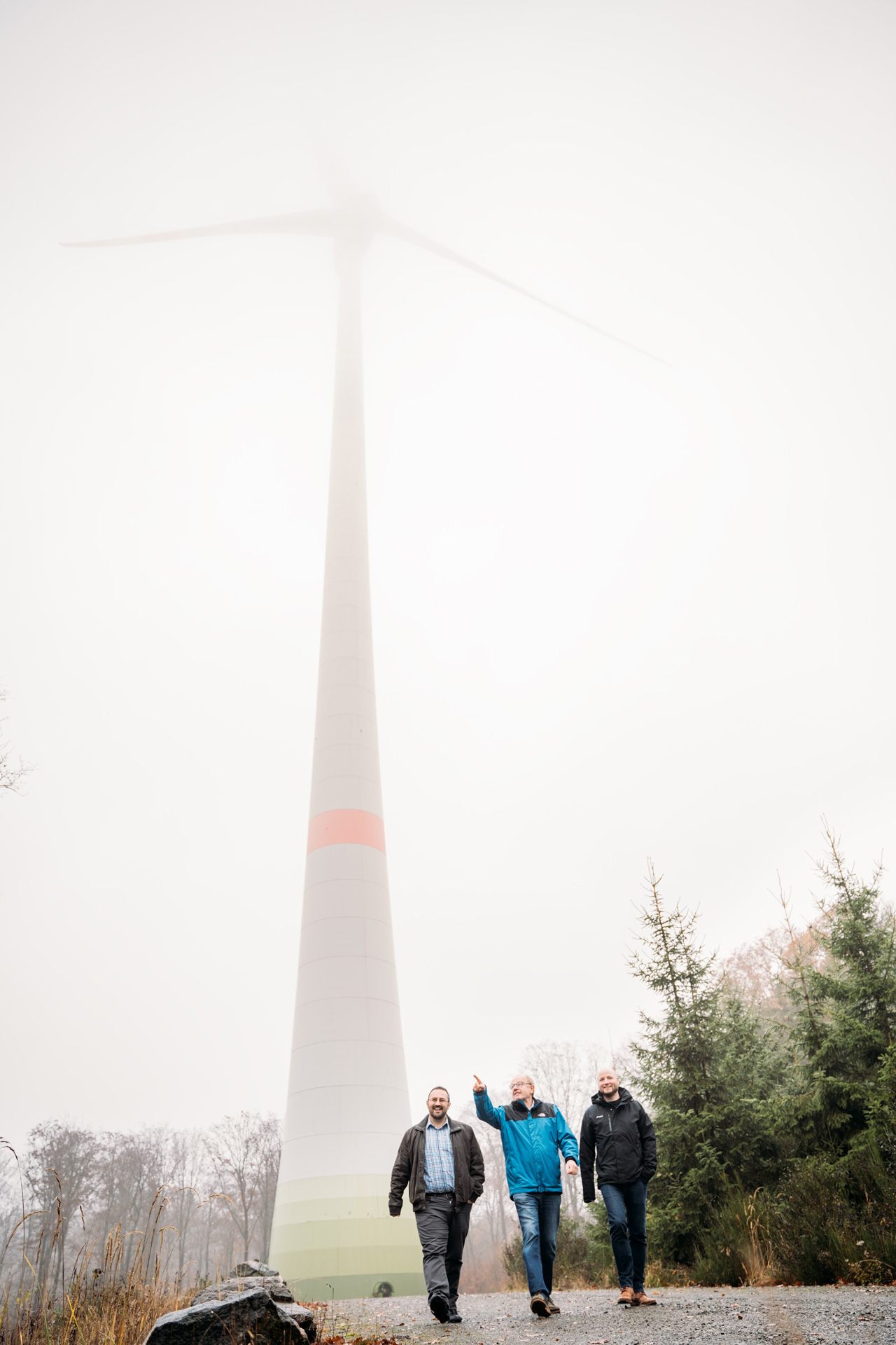Drei Männer gehen auf einem Weg neben einem Windrad, umgeben von Nebel und Bäumen.