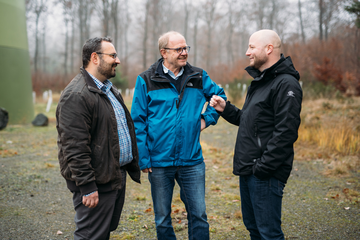 Drei Männer stehen im Freien, lächeln und unterhalten sich, umgeben von unscharfem Hintergrund von Bäumen und Grün.