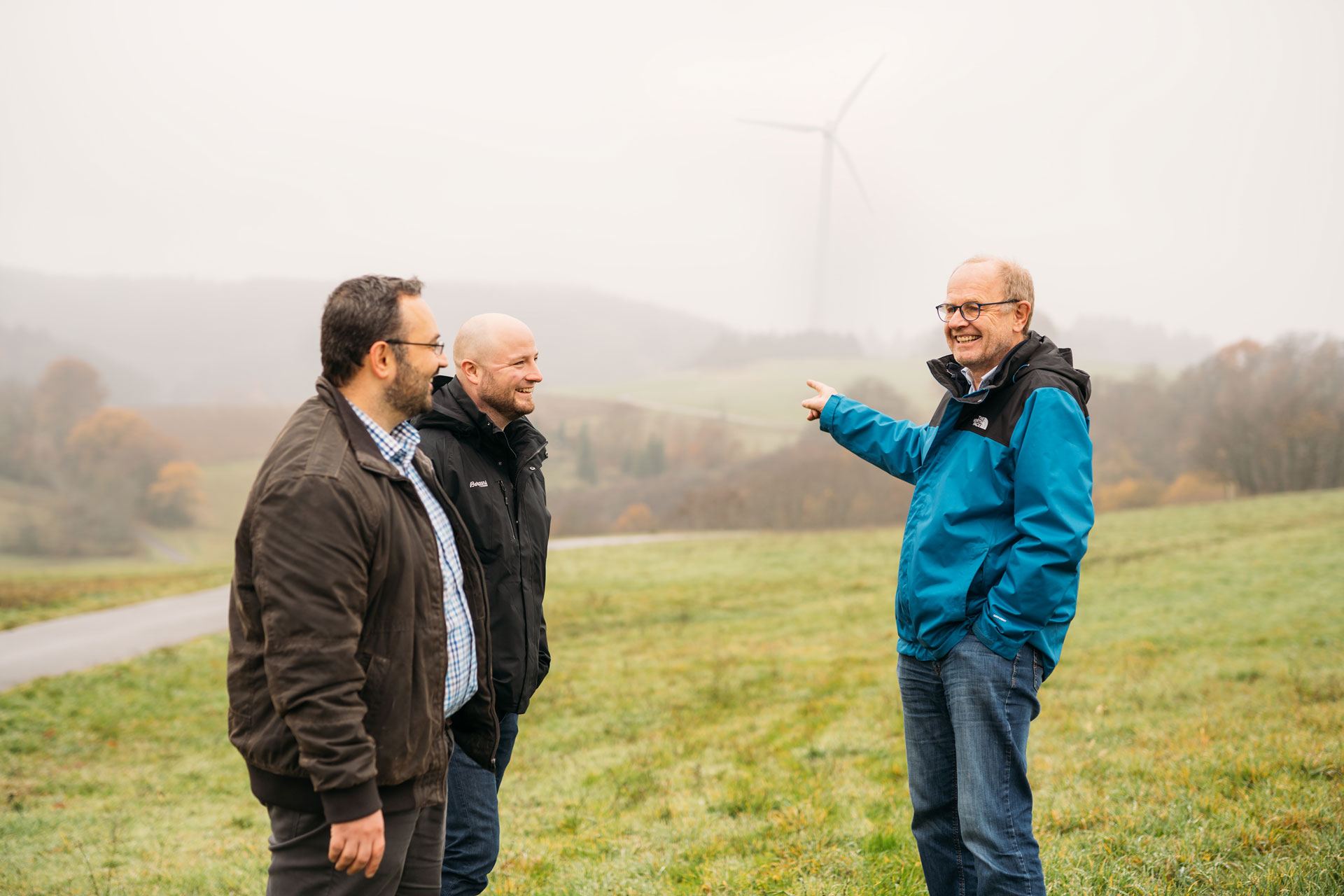Drei Männer stehen im Freien, auf einem sanften Hügel mit Windrädern im Hintergrund, in einer nebligen Landschaft.