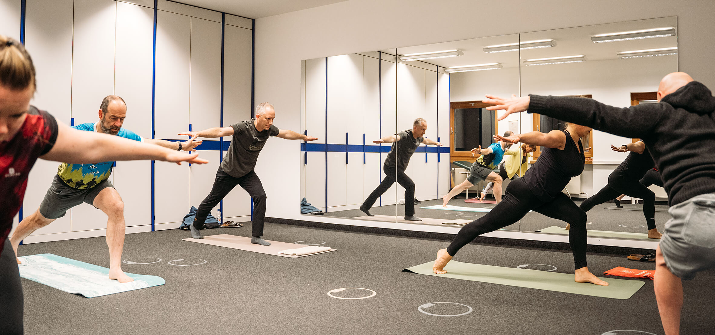 Mehrere Personen praktizieren Yoga in einem hellen Raum mit Spiegeln. Jeder ist auf einer eigenen Matte.
