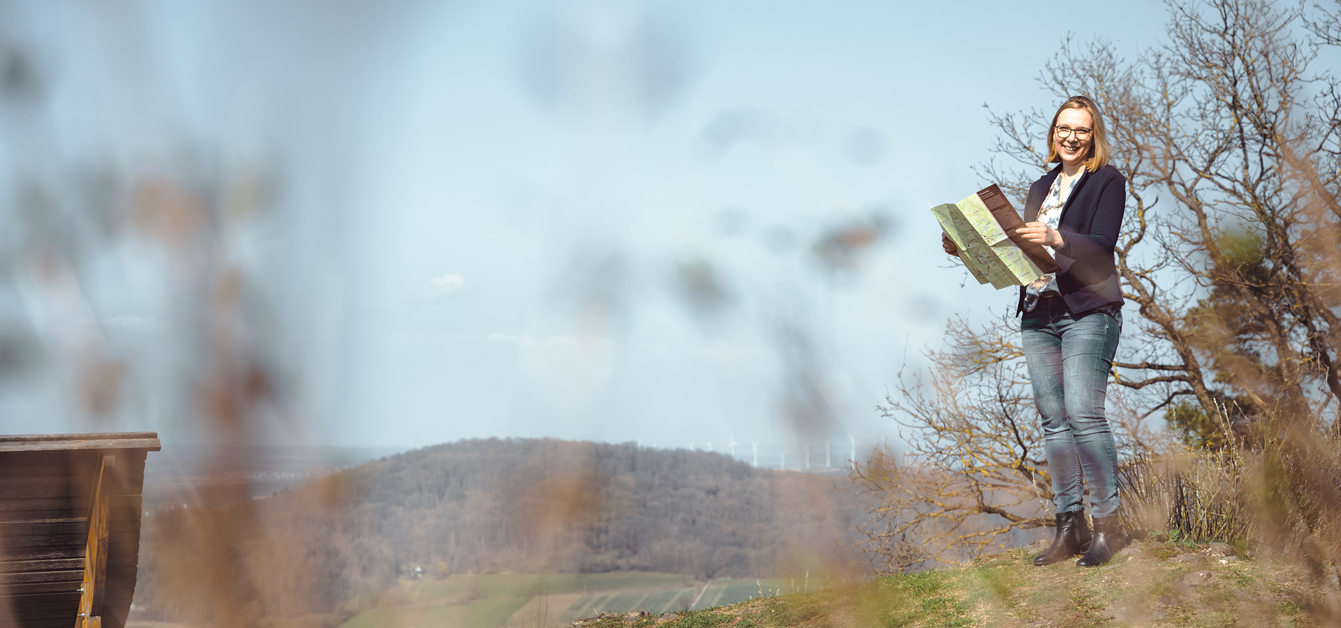 Eine lächelnde Frau steht mit einer Karte in der Hand auf einem Hügel, umgeben von Bäumen und einer weitläufigen Landschaft.