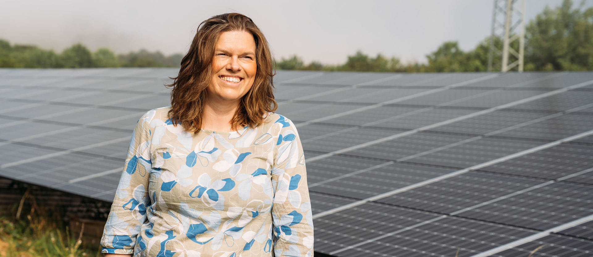 Frau mit lockigem Haar steht lächelnd vor einer Solarflächenanlage an einem bewölkten Tag.