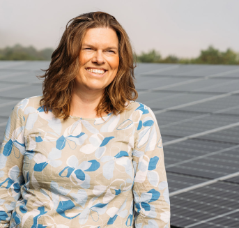 Frau mit lockigem Haar und Blumenmuster-Bluse lächelt vor Solarpanelen im Freien.