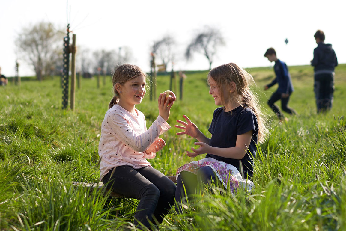 Zwei Kinder sitzen im Gras und teilen einen Apfel, während andere Kinder im Hintergrund spielen.