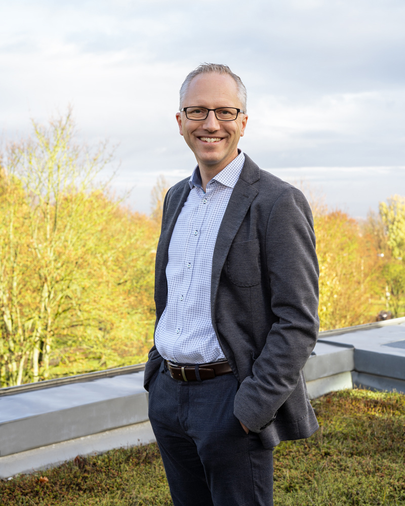 Lächelnder Mann mit Brille im Anzug, stehend auf einem begrünten Dach bei Tageslicht, mit Bäumen im Hintergrund.