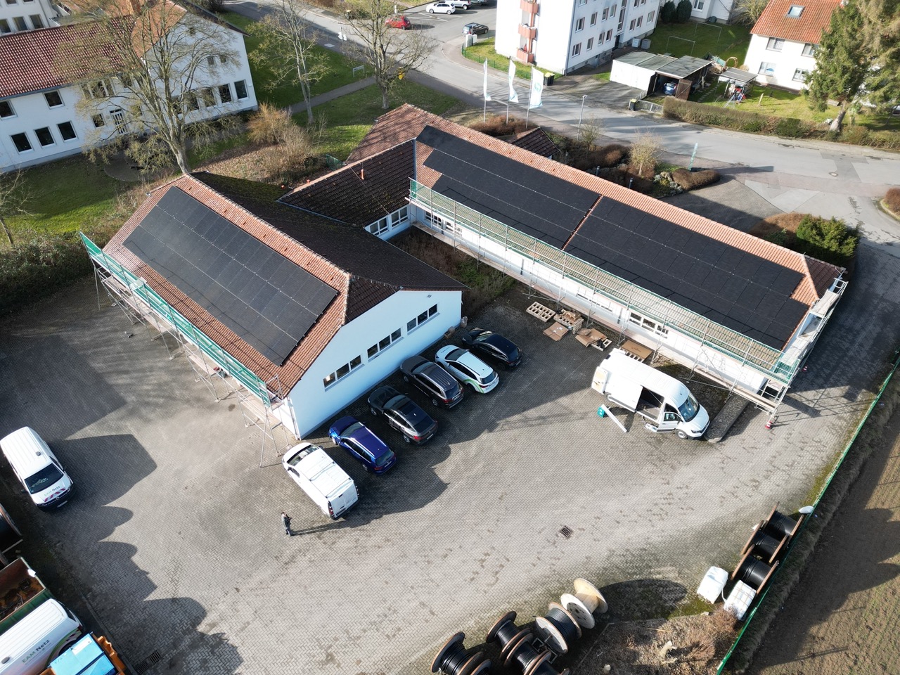 Luftaufnahme eines Gebäudes mit Solarpanelen auf dem Dach, umgeben von Parkplätzen und Bäumen.