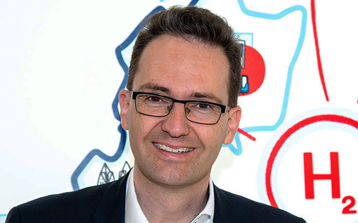 Porträt eines Mannes mit Brille, der lächelt, vor einer bunten, schematischen Hintergrundgrafik.