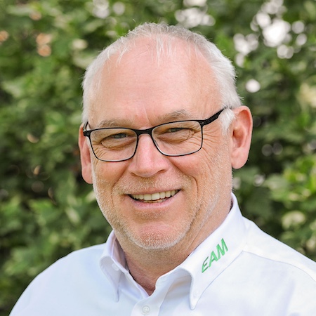 Lächelnder Mann mit Brille und weißem Hemd, das ein grünes Logo trägt, vor einer grünen Pflanzenkulisse.