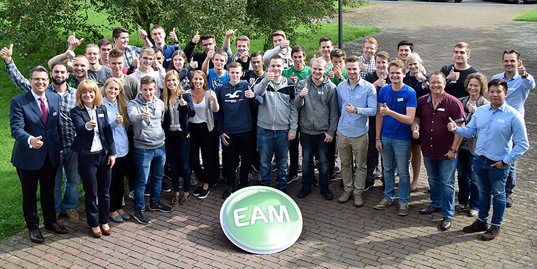 Gruppenshot von Menschen, die zusammen stehen und Daumen hoch zeigen, vor einer grünen Wiese mit dem EAM-Logo im Vordergrund.