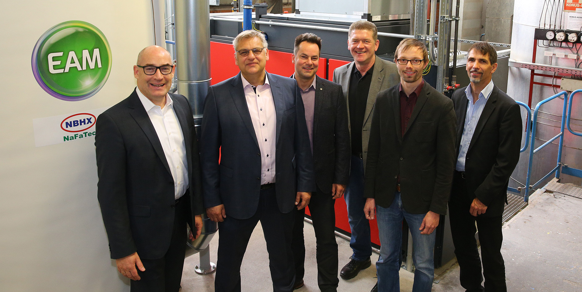 Gruppenfoto von sechs Personen vor einer Maschinenanlage, mit EAM- und NBHX-Logos im Hintergrund.