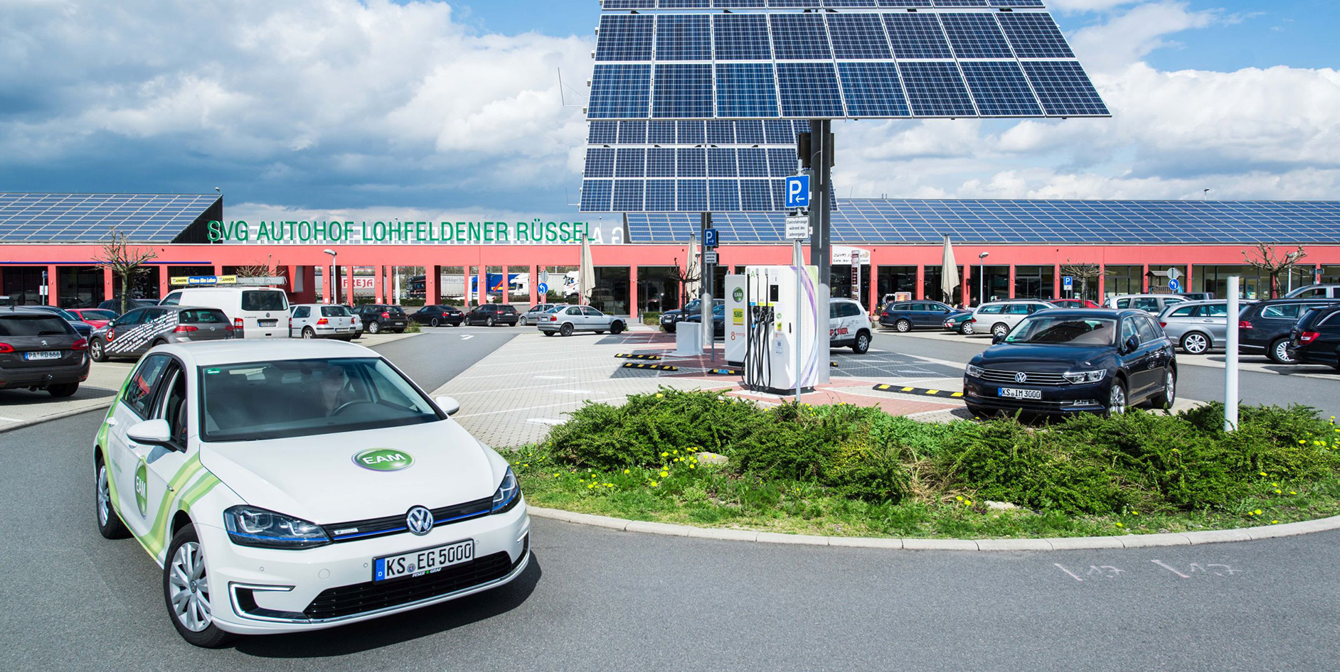 Ein Volkswagen E-Auto parkt an einer Ladestation in einem modernen Autohof mit Solarpanelen im Hintergrund.