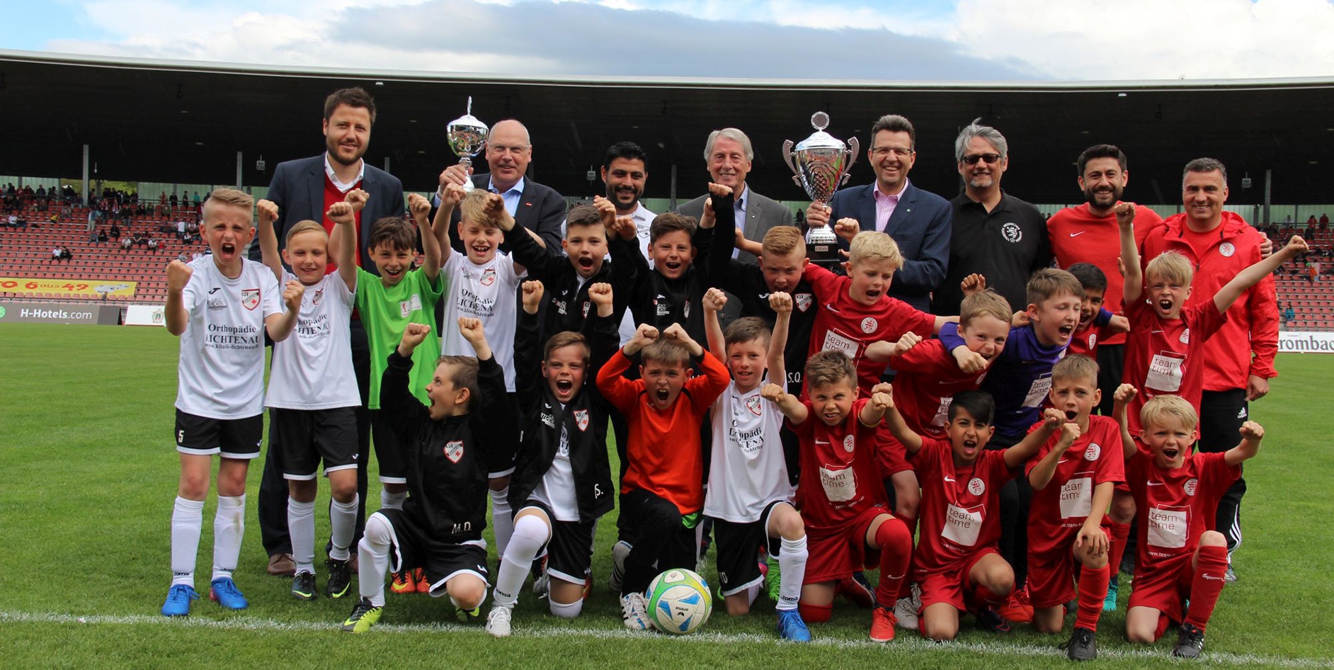 Kinderfußballmannschaft feiert im Stadion mit Pokalen, umgeben von Trainern und Betreuern, in einer jubelnden Pose.