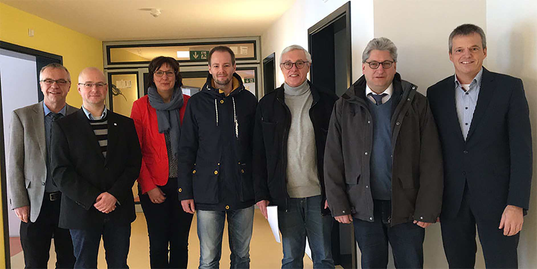 Gruppe von sieben Personen in einem Flur, lächelnd, in formeller Kleidung. Hintergrund mit gelben und weißen Wänden.