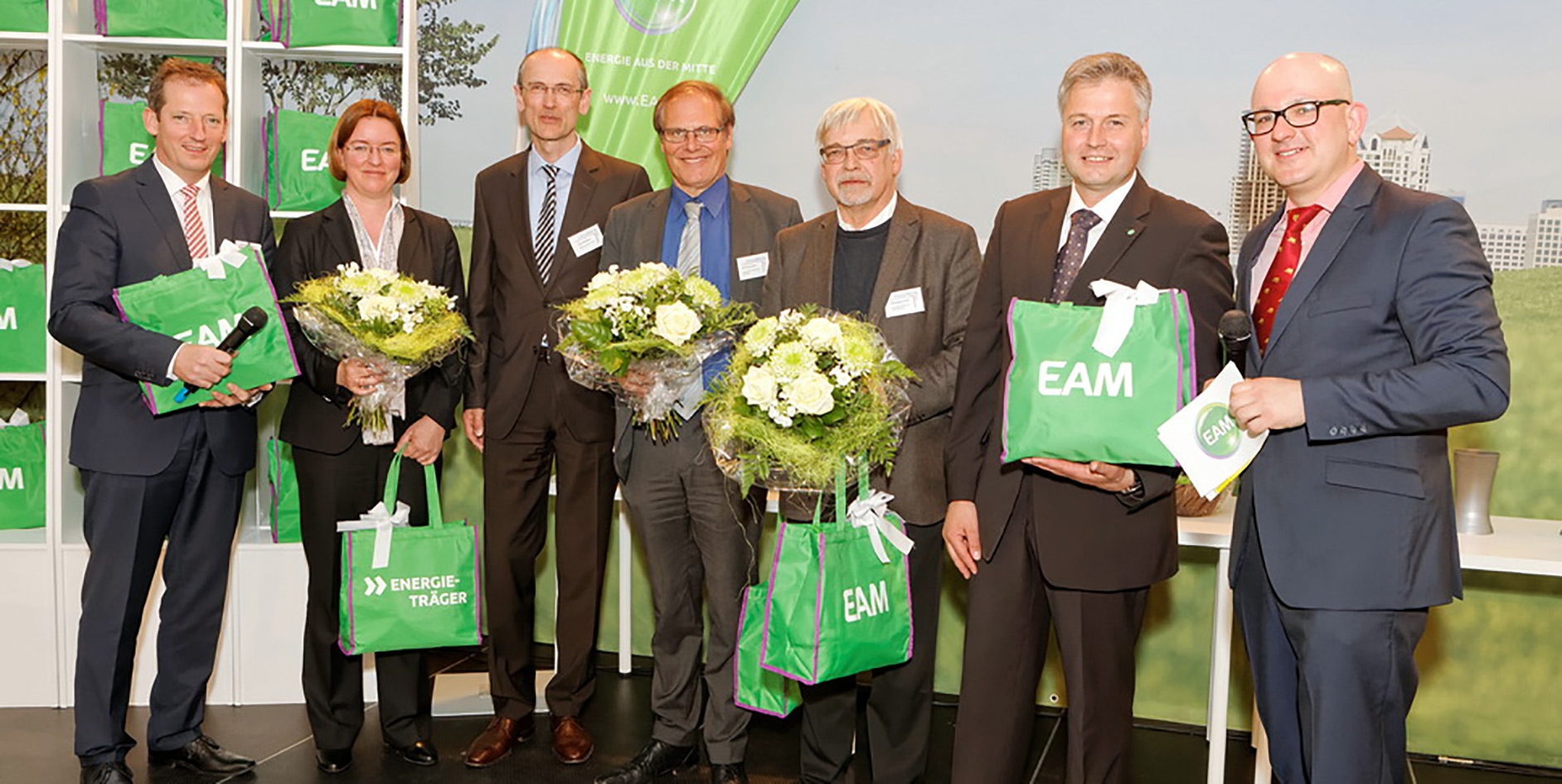 Gruppenfoto von sieben Personen bei einer Veranstaltung, alle halten Blumensträuße und Taschen mit EAM-Logo.