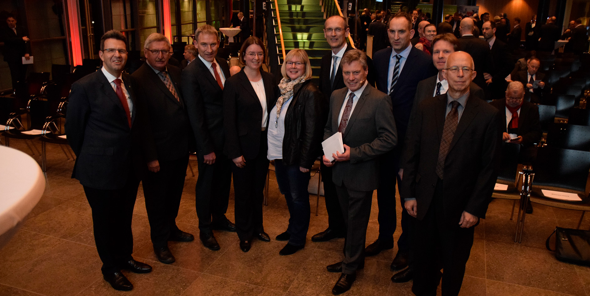 Eine Gruppe von zehn Personen posiert in einem modernen Veranstaltungsraum. Es gibt Stühle und Beleuchtung im Hintergrund.