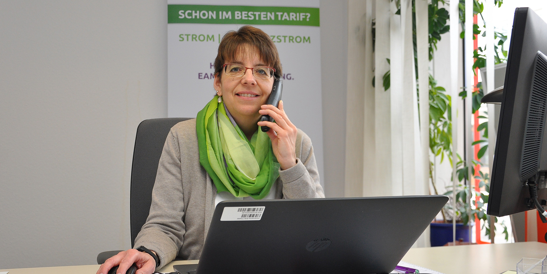 Frau am Telefon im Büro, grüner Schal, am Schreibtisch mit Laptop und Pflanzen im Hintergrund.