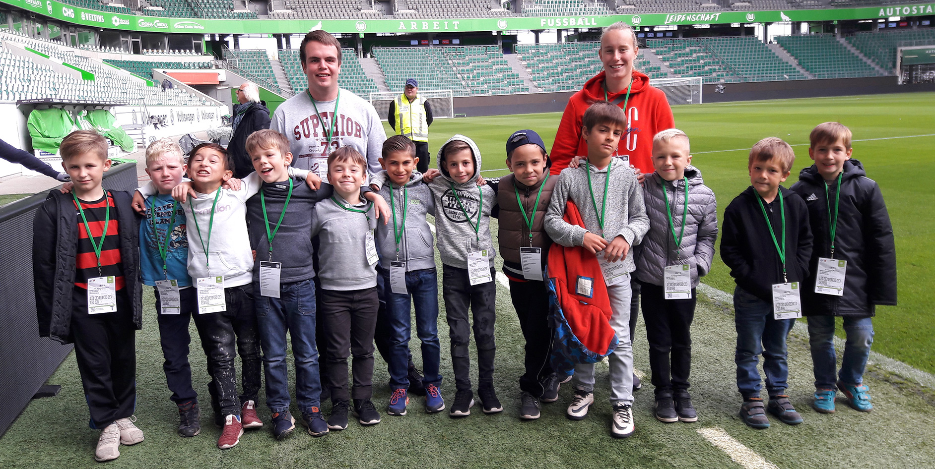 Gruppe von zwölf Kindern mit Lanyards, die auf einem Fußballfeld stehen, mit zwei Erwachsenen im Hintergrund.