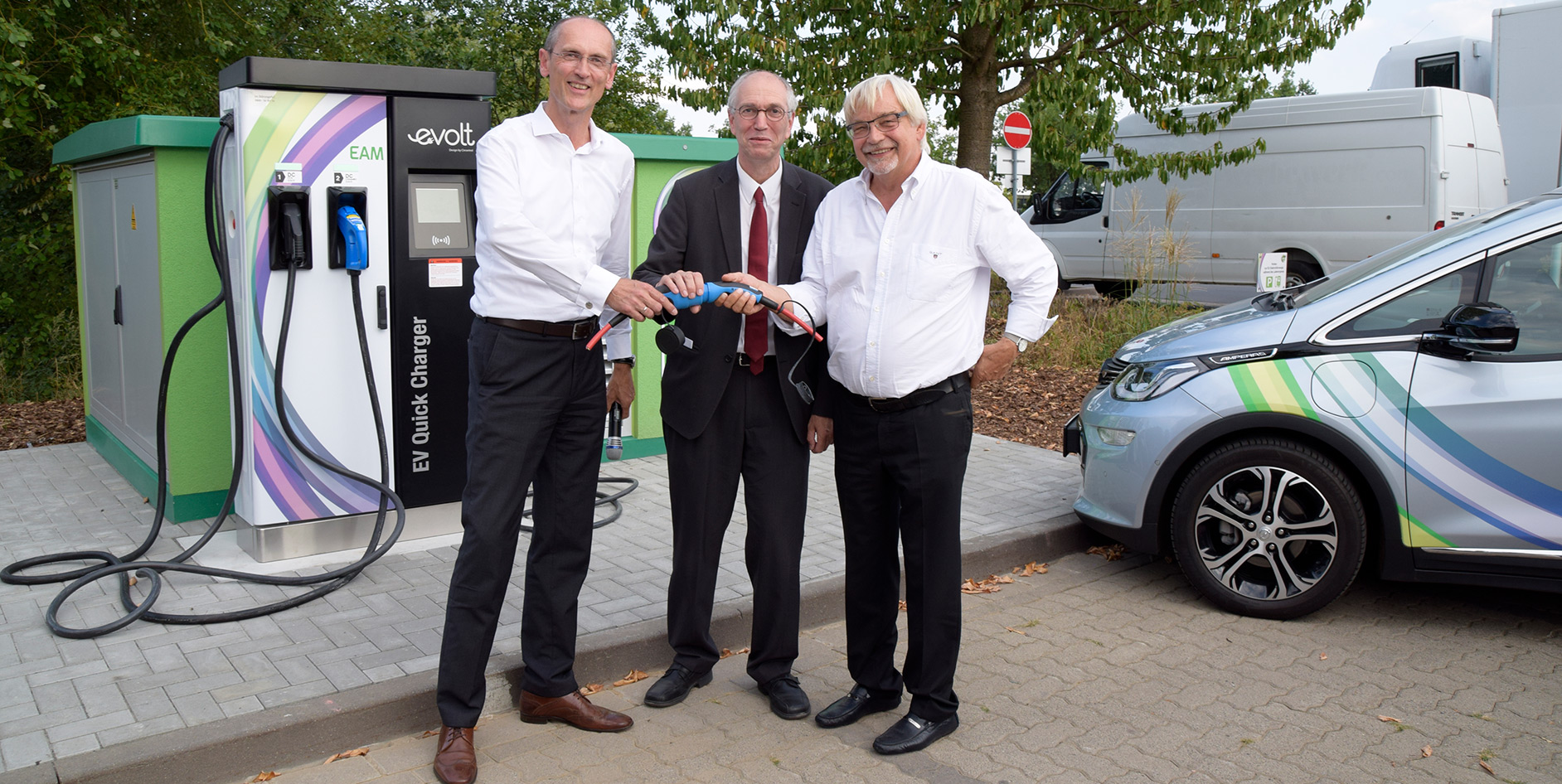Drei Personen stehen an einer Elektro-Ladestation und halten ein Ladekabel in der Hand. Im Hintergrund ist ein Elektrofahrzeug zu sehen.