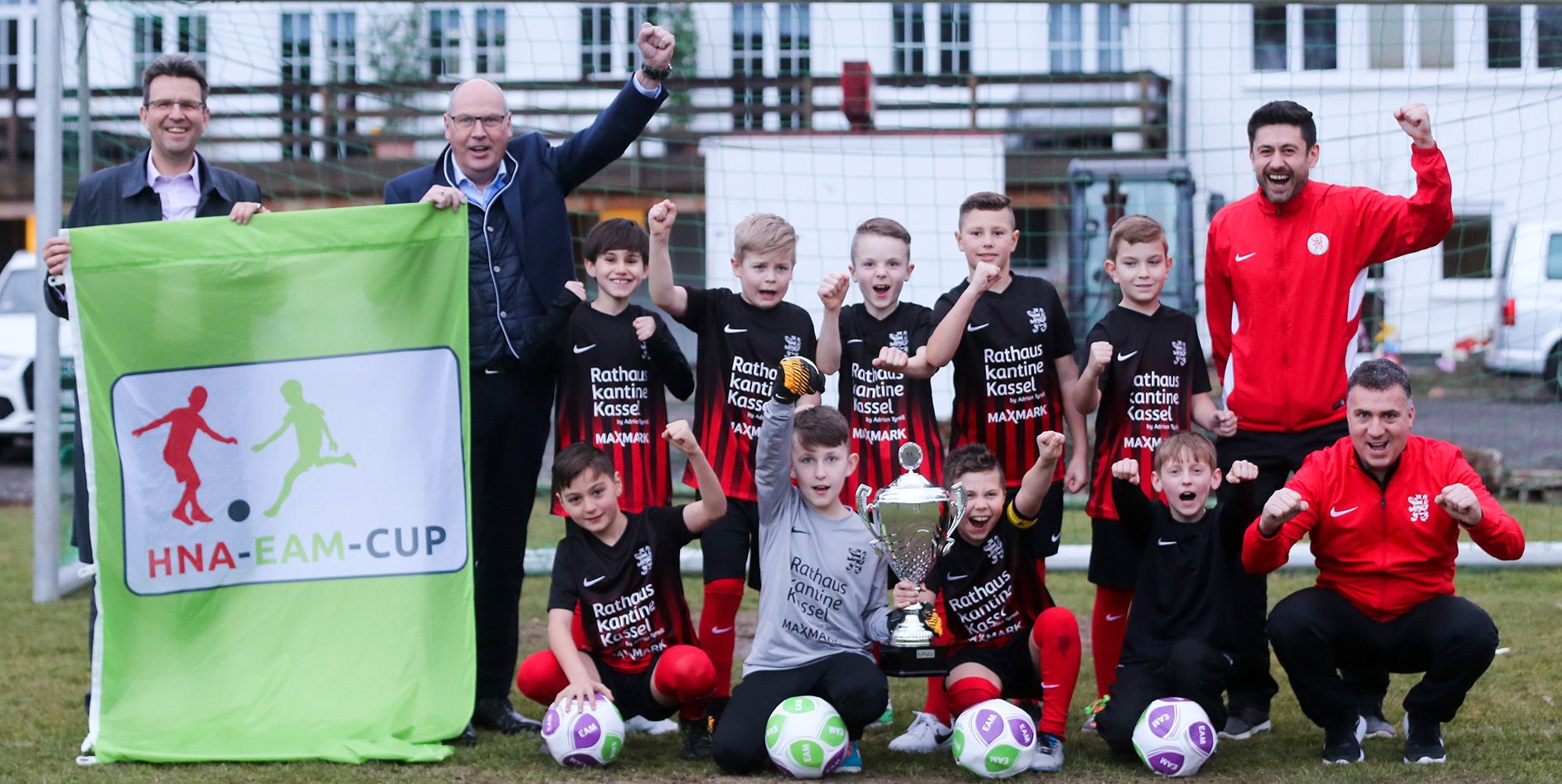 Jubelnde Kinderfußballmannschaft mit Pokal und Banner des HNA-EAM-Cups auf dem Spielfeld.