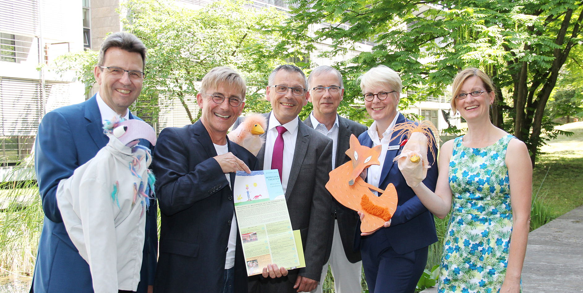 Gruppenfoto von sechs Personen im Freien, die Handpuppen und ein Plakat halten, umgeben von Bäumen und Pflanzen.