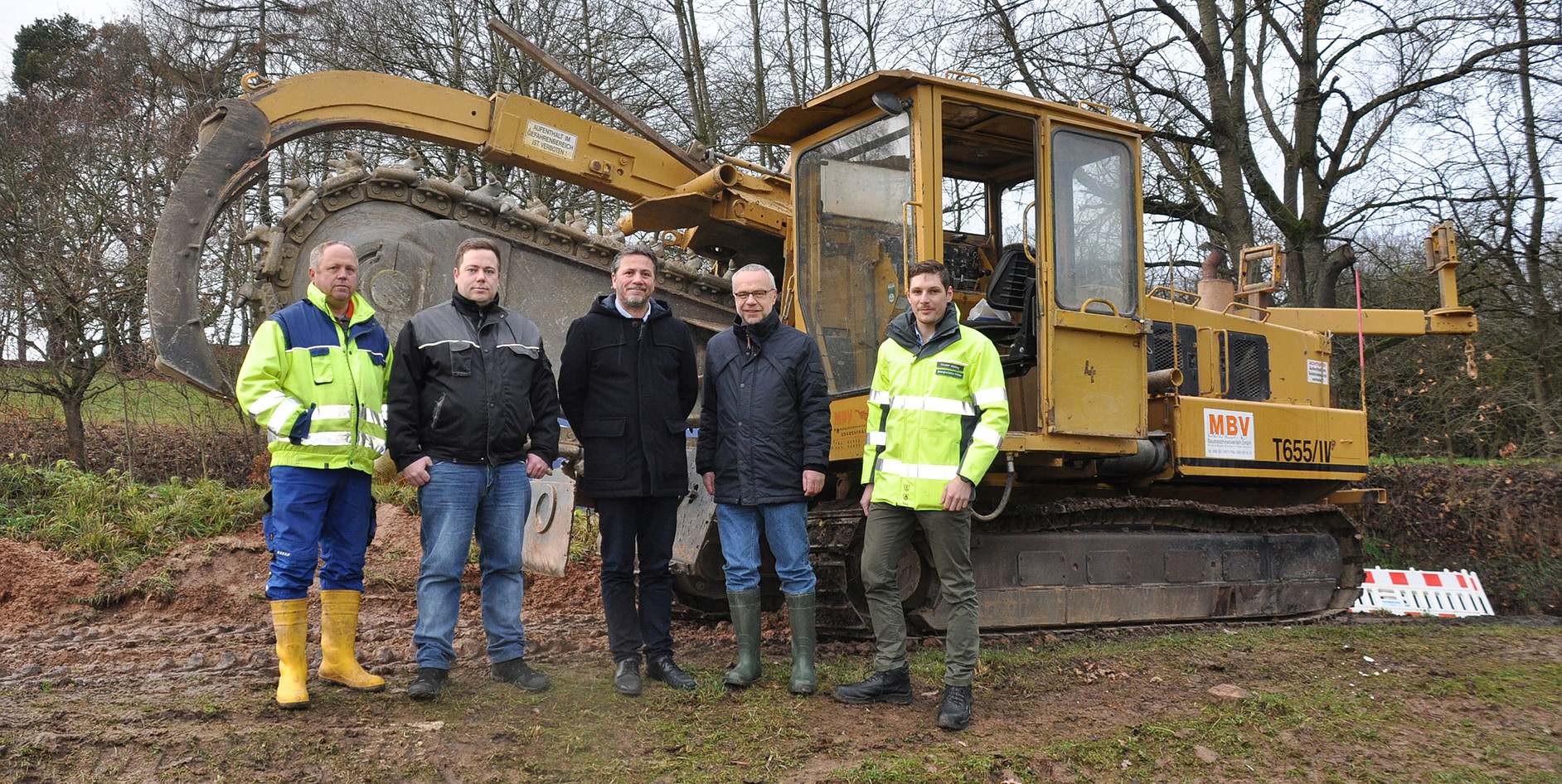 Fünf Personen stehen vor einem gelben Baumaschinenfahrzeug in einer ländlichen Umgebung.