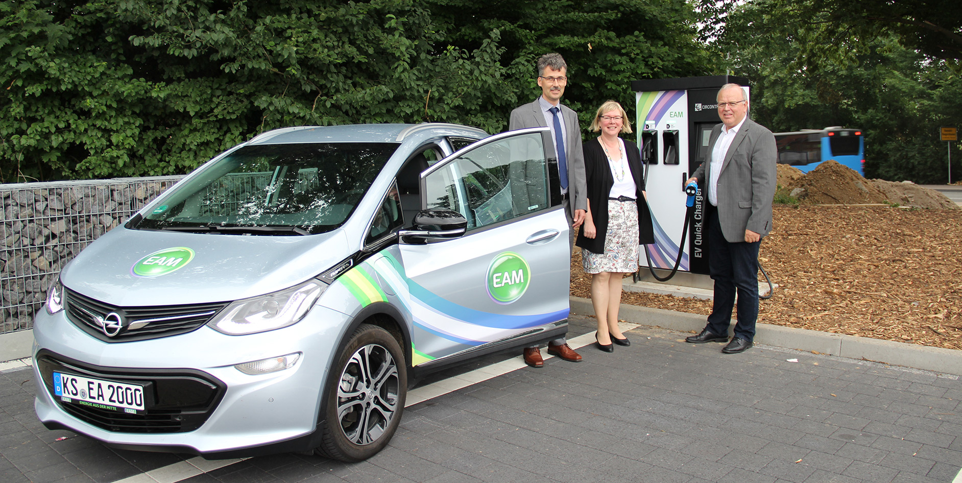 Drei Personen stehen neben einem Elektrofahrzeug an einer Ladestation mit EAM-Logo im Hintergrund.