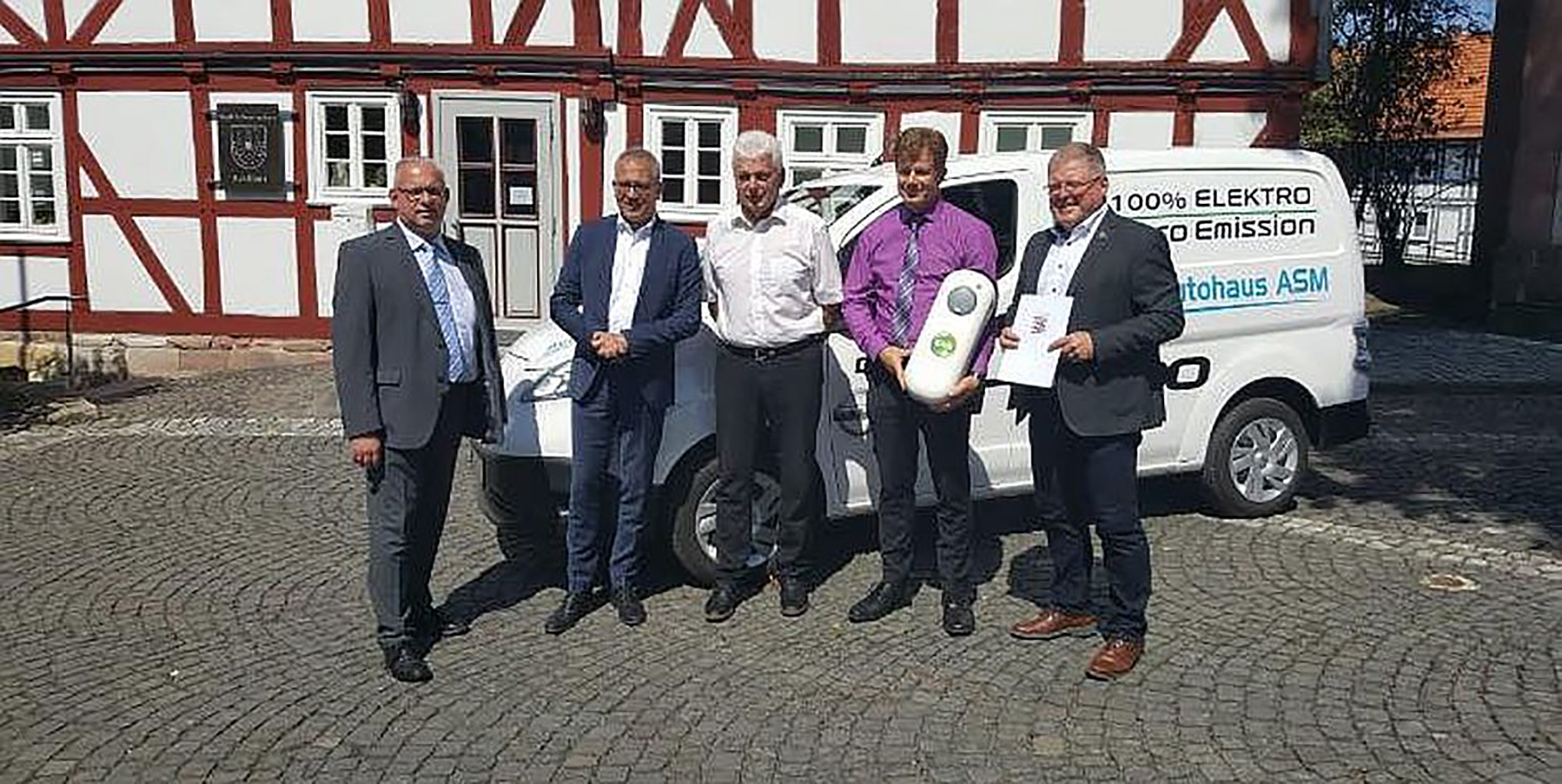 Fünf Personen stehen vor einem Elektrofahrzeug vor einem Fachwerkhaus, sie halten Dokumente und ein Objekt in der Hand.