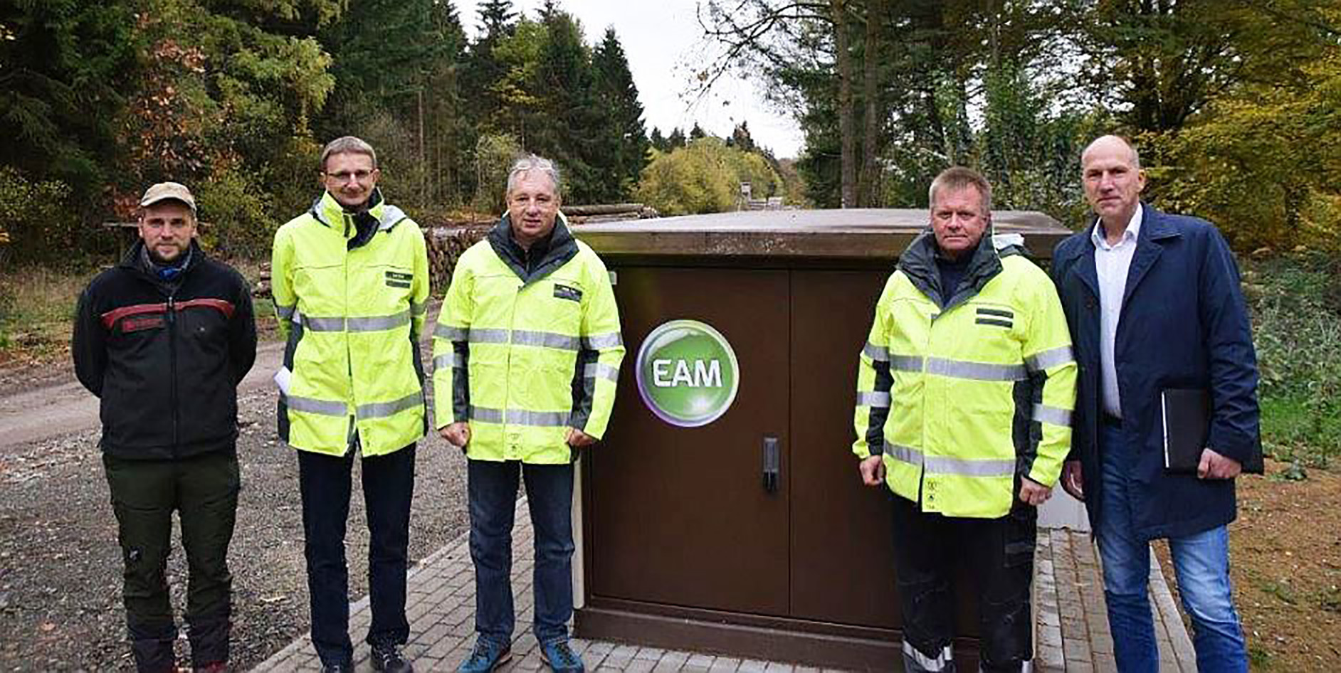 Fünf Personen in Sicherheitskleidung stehen vor einem Technikschrank im Wald, umgeben von Bäumen und herbstlichen Farben.